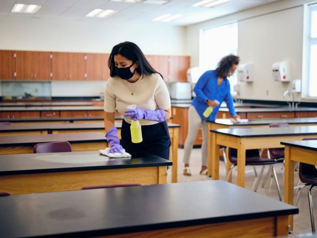 Cleaning staff sanitizing classroom desks with disinfectant spray for a hygienic learning environment.
