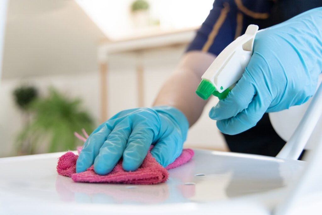 Gloved hand spraying disinfectant while wiping a hard surface with a microfiber cloth.