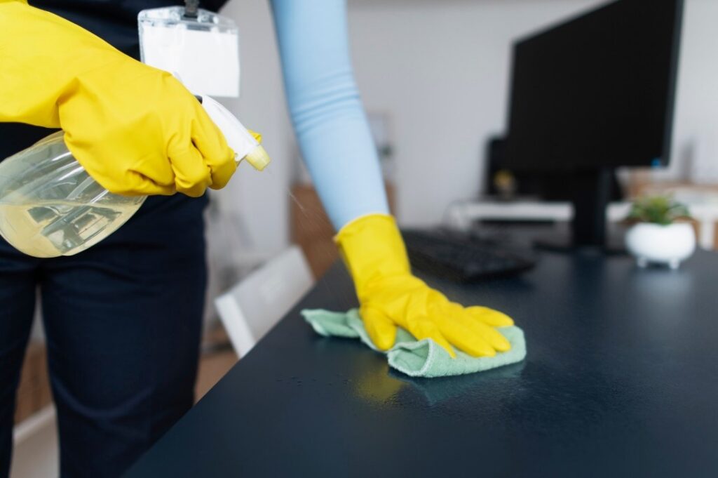 Professional cleaner disinfecting an office desk using spray cleaner and a microfiber cloth.